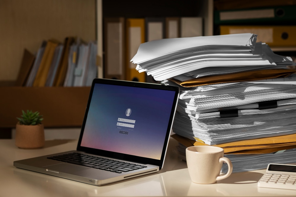 Stack of documents beside an open laptop on a desk.