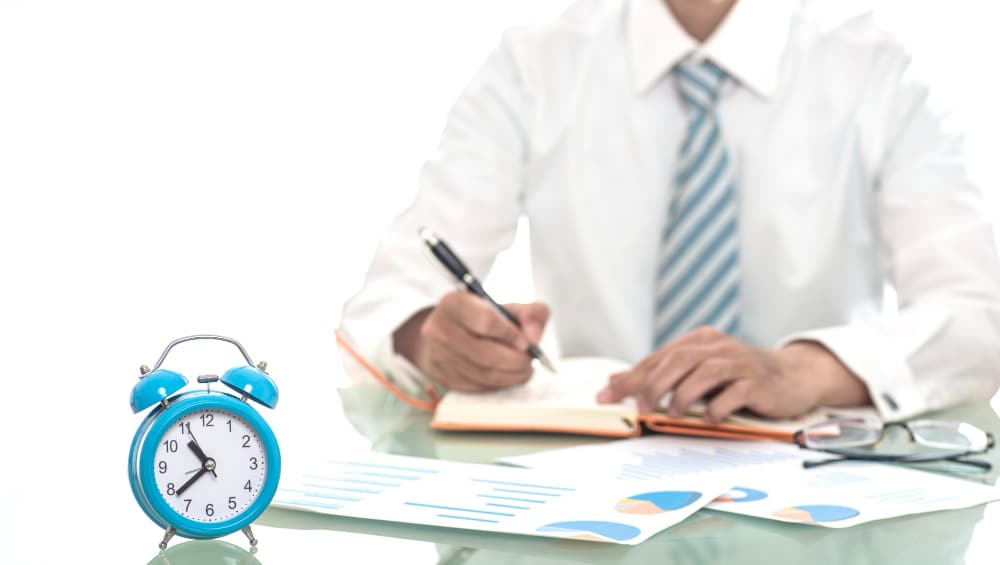 Young professional taking notes on paper with a clock placed nearby on the table.