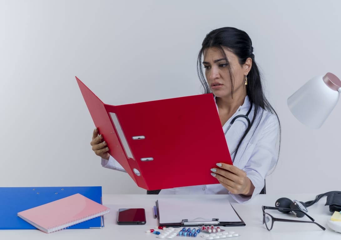 Concerned young female doctor sitting at desk with medical tools, reviewing a patient folder